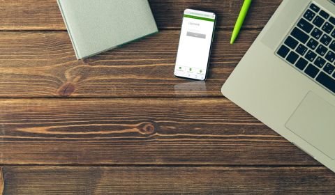 drewniane deski biurka, w prawym rogu widać część laptopa, zielony długopis, kalendarz i ekran telefonu komórkowego