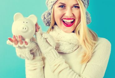 kobieta ubrana w szarą zimową czapkę biały szal i biały sweter trzyma w ręku świnkę skarbonkę